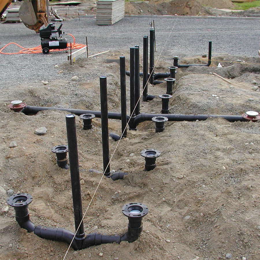 Underground plumbing rough-in with black drain stubs rising in organized rows from a gravel base in an open trench.